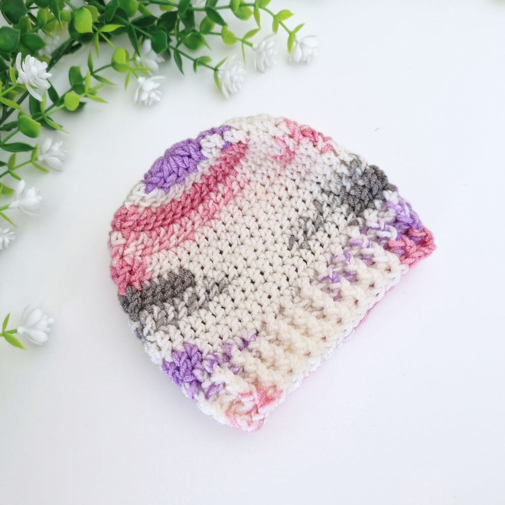 A handmade crocheted beanie hat made from variegated yarn in shades of pink, purple, gray, and cream, styled on a white surface with green leaves and small white flowers in the corner.