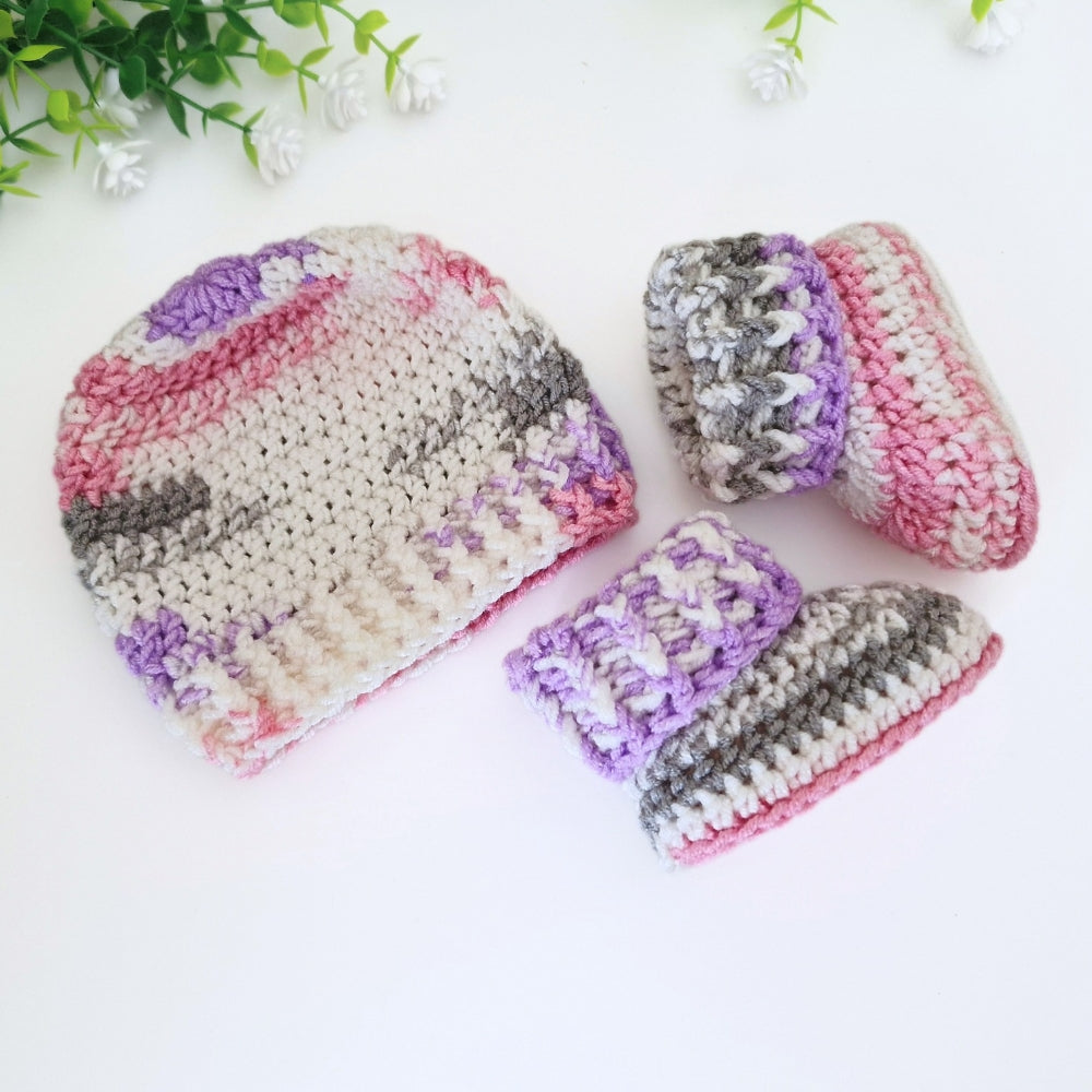 A handmade crochet baby hat and matching booties in a variegated yarn of pink, purple, gray, and white, arranged on a white background with decorative green leaves in the corner.