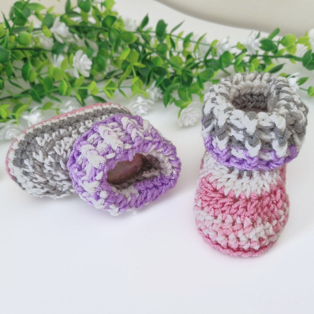 A pair of handmade crocheted baby booties in pastel colors, including gray, purple, white, and pink, arranged on a white surface with decorative green leaves in the background.
