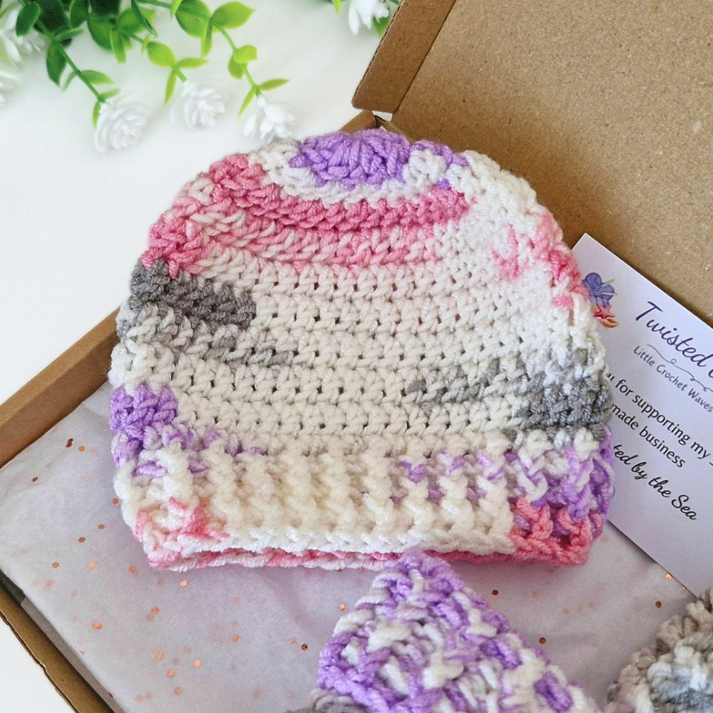 A handmade crocheted baby hat in a cream, pink, purple, and gray striped pattern, displayed inside a kraft paper gift box with a brand card.