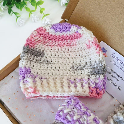 A handmade crocheted baby hat in a cream, pink, purple, and gray striped pattern, displayed inside a kraft paper gift box with a brand card.