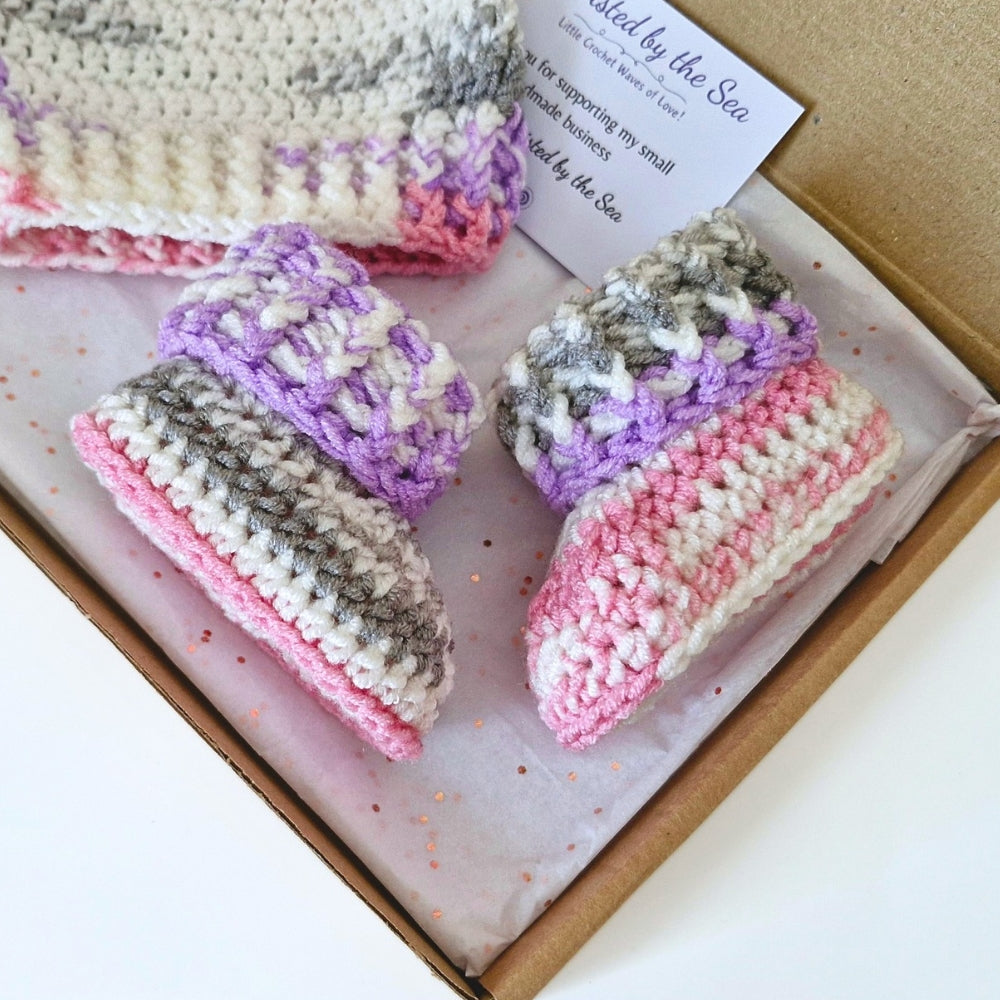 A handmade crochet baby bootie set with a matching hat, displayed inside a gift box. The booties feature stripes of pink, cream, and gray, with textured purple and gray details.