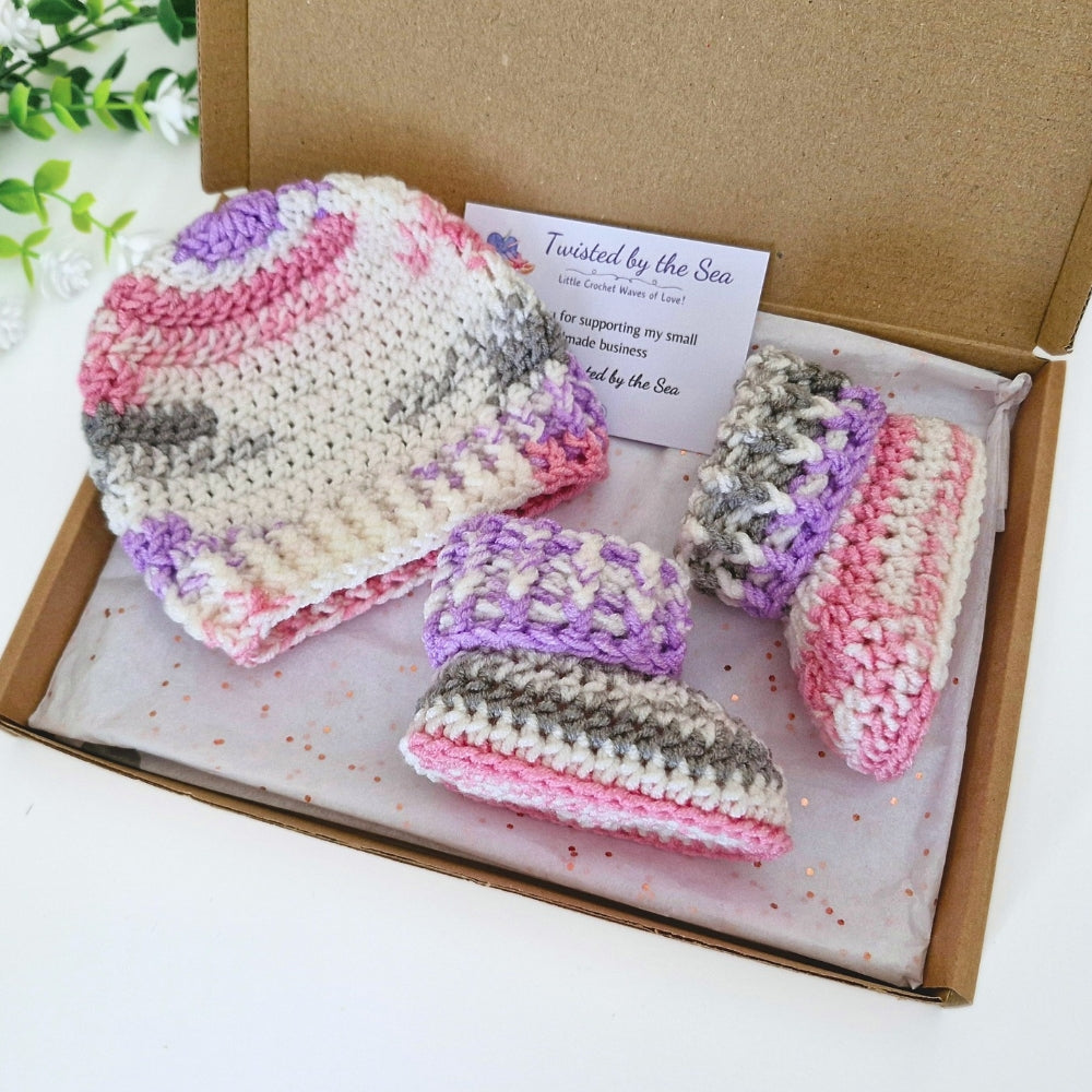 A handmade crochet baby hat and matching booties set in a brown cardboard gift box. The crochet set features stripes of pink, purple, gray, and cream.