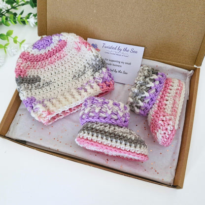 A handmade crochet baby hat and matching booties set in a brown cardboard gift box. The crochet set features stripes of pink, purple, gray, and cream.