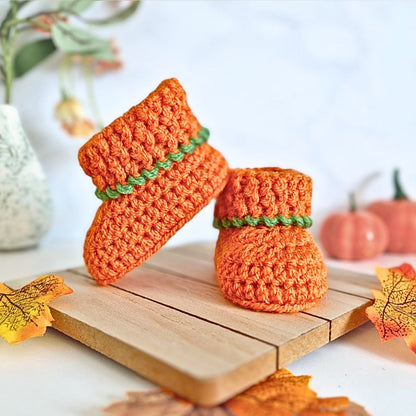 Two pumpkin orange crochet pumpkins with green stems on a wooden surface with autumn leaves.