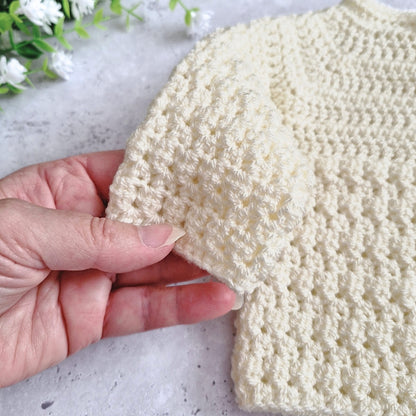 A close-up of a hand holding the sleeve of a cream-colored, textured crochet knit garment, showing the fabric detail against a light gray stone background with white flowers.