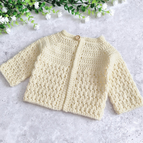 A cream-coloured, long-sleeve crochet cardigan with a single wooden button closure, laid flat on a light gray textured surface with decorative green and white artificial flowers in the top corner.