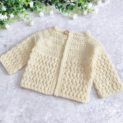 A cream-coloured, long-sleeve crochet cardigan with a single wooden button closure, laid flat on a light gray textured surface with decorative green and white artificial flowers in the top corner.