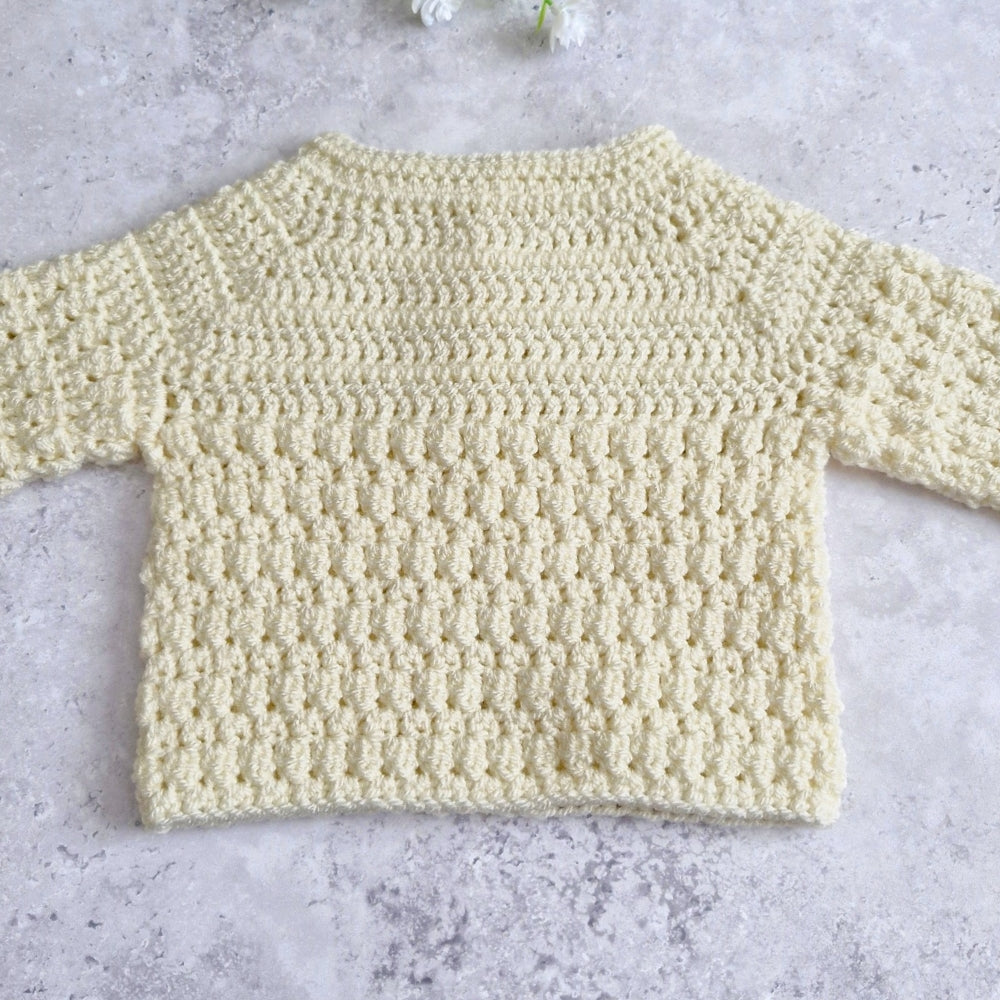 Tge back view of a pale cream crocheted long-sleeve top laid flat on a light gray textured surface.