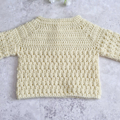 Tge back view of a pale cream crocheted long-sleeve top laid flat on a light gray textured surface.
