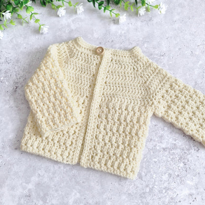 A cream-coloured, hand-crocheted baby cardigan with long sleeves and a single wooden button, laid flat on a light gray textured surface with small white flowers in the corner.