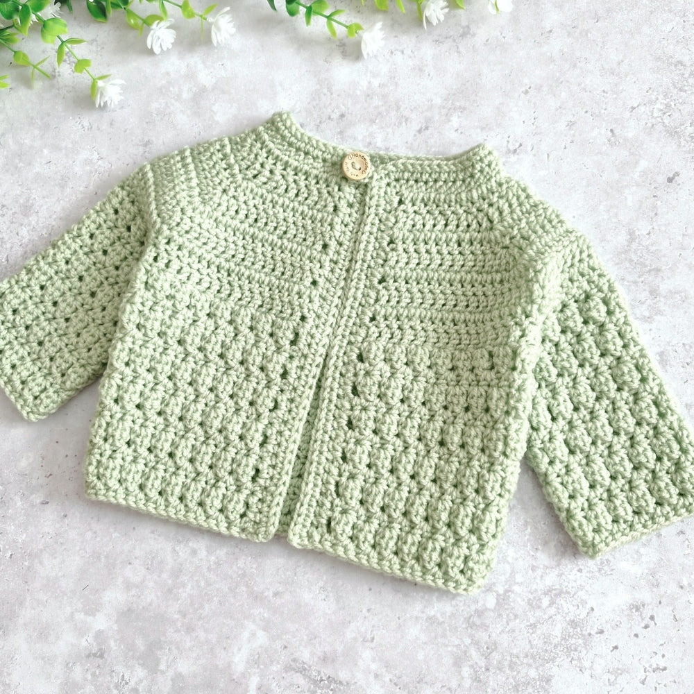 A light tea green crocheted cardigan sweater with long sleeves and a single wooden button, laid flat on a light gray textured surface with small white flowers in the corner.