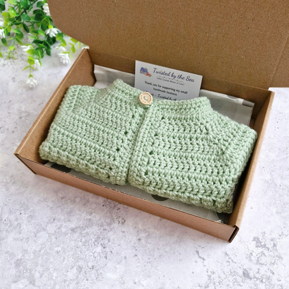 A light green crocheted baby cardigan with a single wooden button, neatly folded inside a brown cardboard gift box with a brand card.