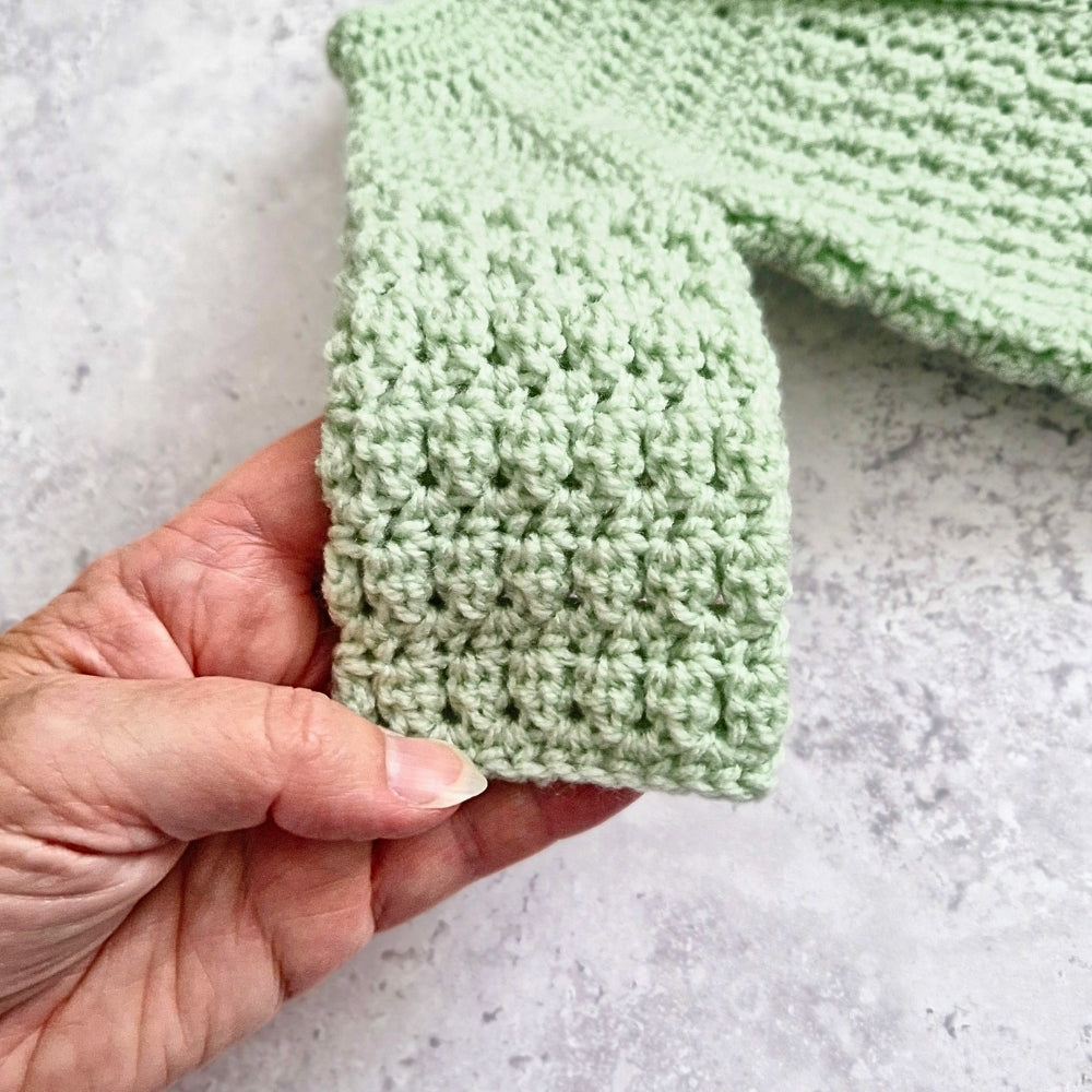 A close-up of a hand holding a light green crocheted sleeve or cuff, showing the textured knit pattern against a light gray stone-like background.