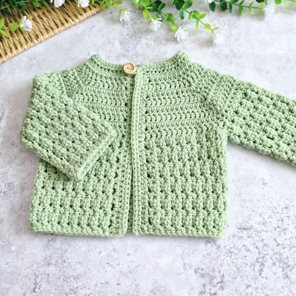 A light green crocheted cardigan sweater with long sleeves and a single wooden button, laid flat on a light gray textured surface with small white flowers and a basket in the background.