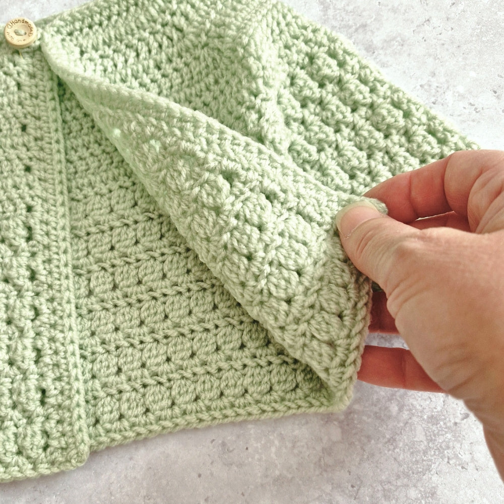 A close-up of a light sage green crocheted textile, likely a baby blanket or scarf, being held by a hand to show the texture. The item features a textured bobble stitch pattern, a ribbed border, and a small wooden button on the left, set against a light gray stone background.