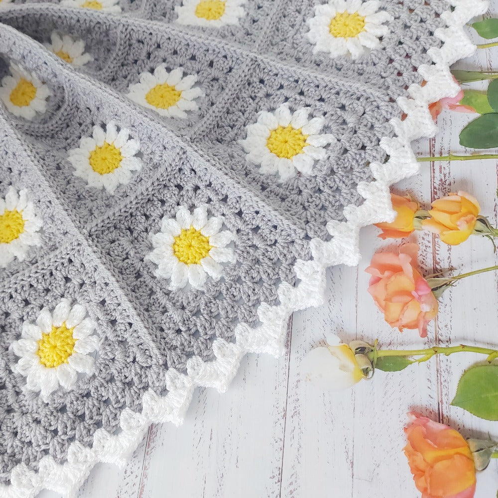 Gray crocheted blanket with white daisy patterns on a wooden surface with flowers.