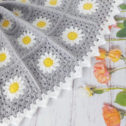 Gray crocheted blanket with white daisy patterns on a wooden surface with flowers.