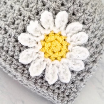 Close-up of a handmade crocheted daisy with white petals and yellow center on gray yarn background