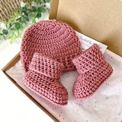 Pink crochet baby hat and booties set on a white box with greenery in the background