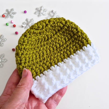 A hand holding a small crocheted beanie hat with an mistletoe green top and a white brim, photographed against a light background with decorative silver snowflakes.