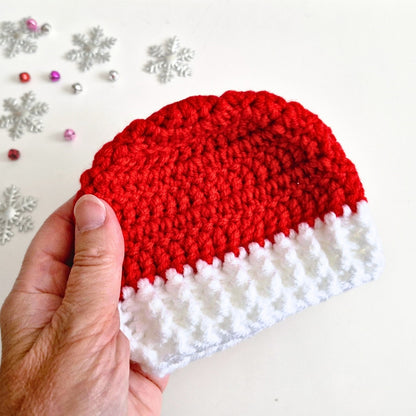 A hand holding a small crocheted Santa hat in red and white, with silver snowflake decorations on a plain light background.