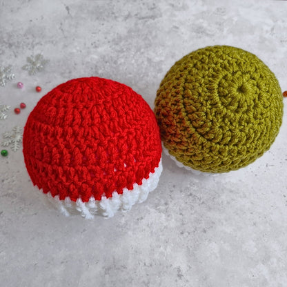 Two crocheted round hats displayed on a light gray textured surface. The hat on the left is red with a white bottom trim, and the hat on the right is mistletoe green with a white bottom trim.