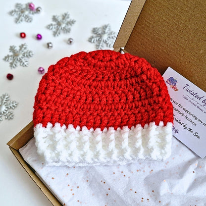 A red and white crocheted Santa hat displayed inside a brown gift box with tissue paper and a branded card. The box is set on a white surface decorated with silver snowflakes and small colorful beads.