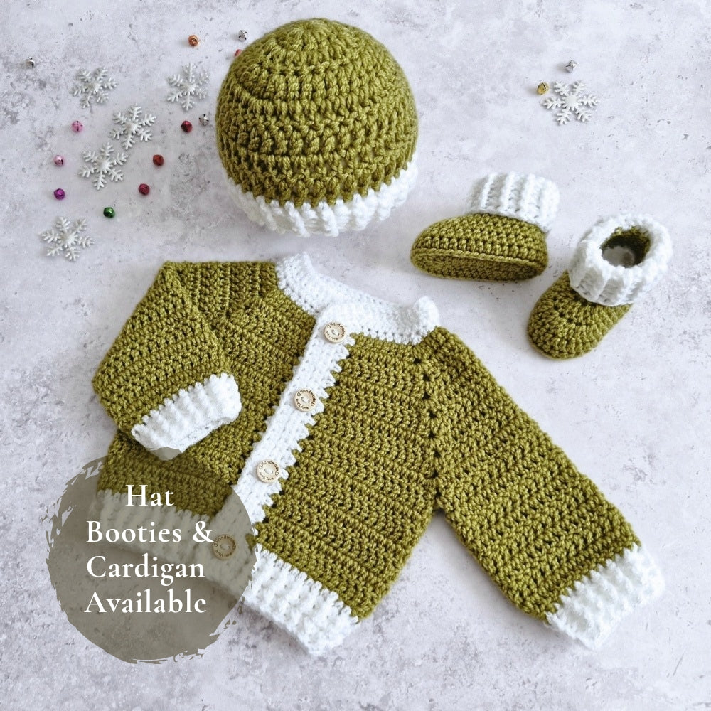 Crocheted baby outfit set laid flat on a light textured surface, including a hat, cardigan, and booties in mistletoe green and white, with small decorative snowflake ornaments and a text badge that says 'Hat Booties & Cardigan Available'.