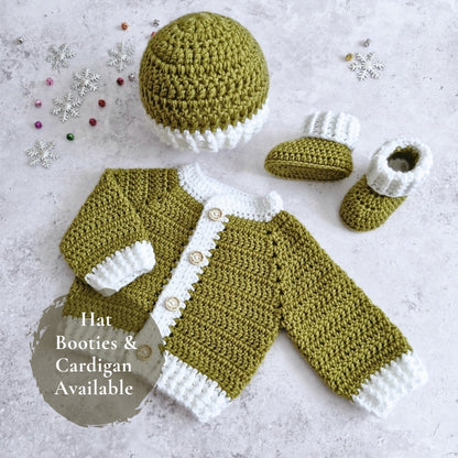 Crocheted baby outfit set laid flat on a light textured surface, including a hat, cardigan, and booties in mistletoe green and white, with small decorative snowflake ornaments and a text badge that says 'Hat Booties & Cardigan Available'.