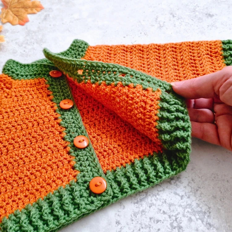 A close-up of a crocheted baby pumpkin coloured cardigan in orange and green, held open by a hand. The cardigan features orange panels bordered by green ribbed trim and four orange buttons.