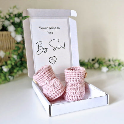 Pink baby booties in a box with a card reading 'You're going to be a Big Sister!' on a white surface with greenery in the background.