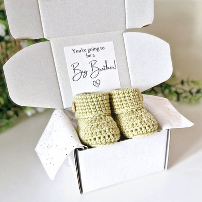 Newborn crochet booties in a box with a card reading 'You're going to be a Big Brother!' on a light background.