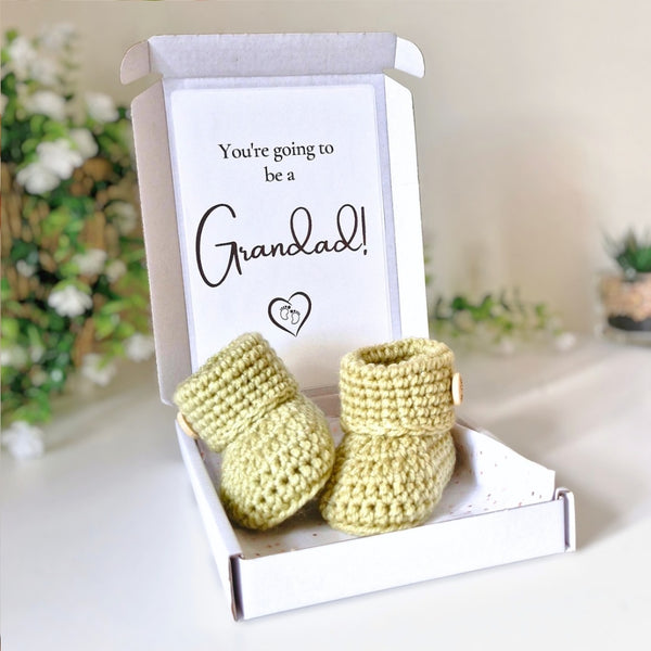Newborn green crocheted booties in a box with a card reading 'You're going to be a Grandad!' on a blurred background.