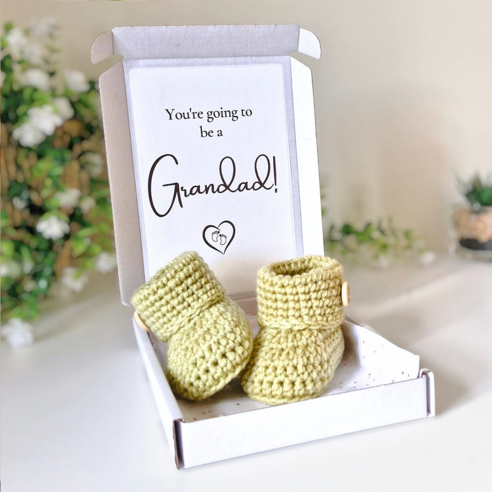 Newborn green crocheted booties in a box with a card reading 'You're going to be a Grandad!' on a blurred background.