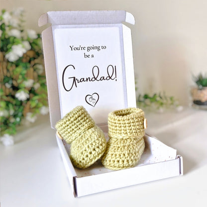 Newborn green crocheted booties in a box with a card reading 'You're going to be a Grandad!' on a blurred background.