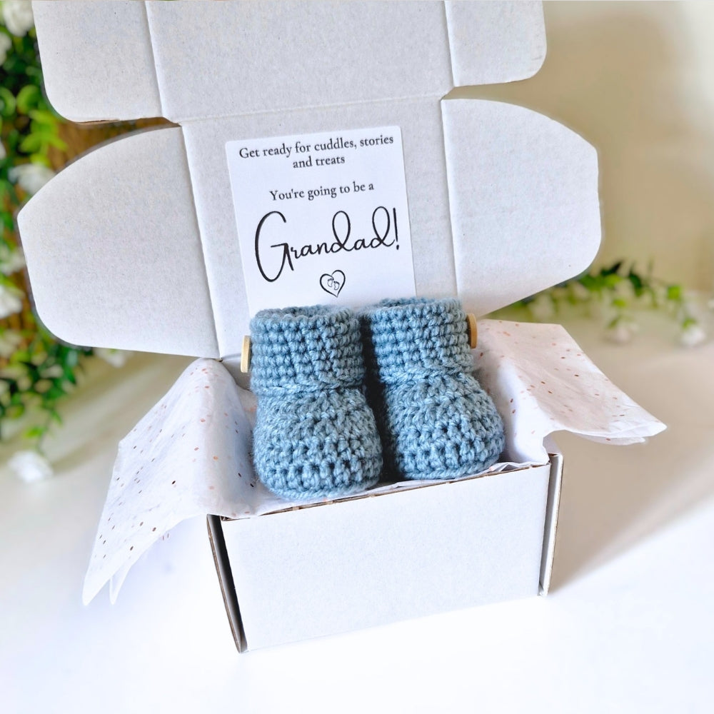 Blue baby booties in a white box with a card inside announcing being a Grandad, set against a blurred floral background.