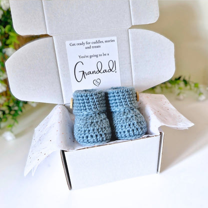 Blue baby booties in a white box with a card inside announcing being a Grandad, set against a blurred floral background.