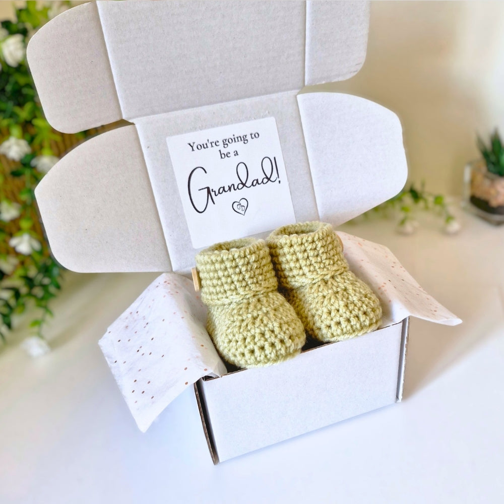 Newborn green crochet baby booties in a box with a card reading 'You're going to be a Grandad!' on a light background.