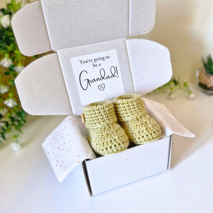 Newborn green crochet baby booties in a box with a card reading 'You're going to be a Grandad!' on a light background.