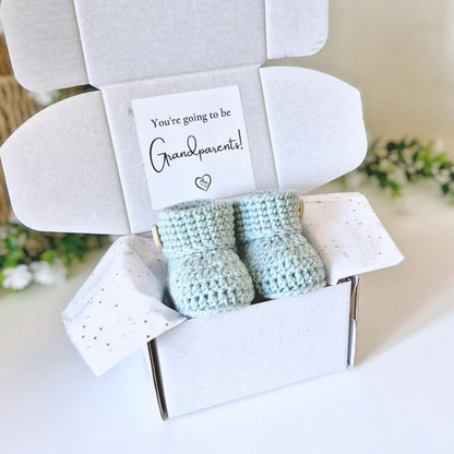 Grandparents to be pregnancy announcement with a Pair of light blue baby booties in a white box with a card inside, set against a blurred background.
