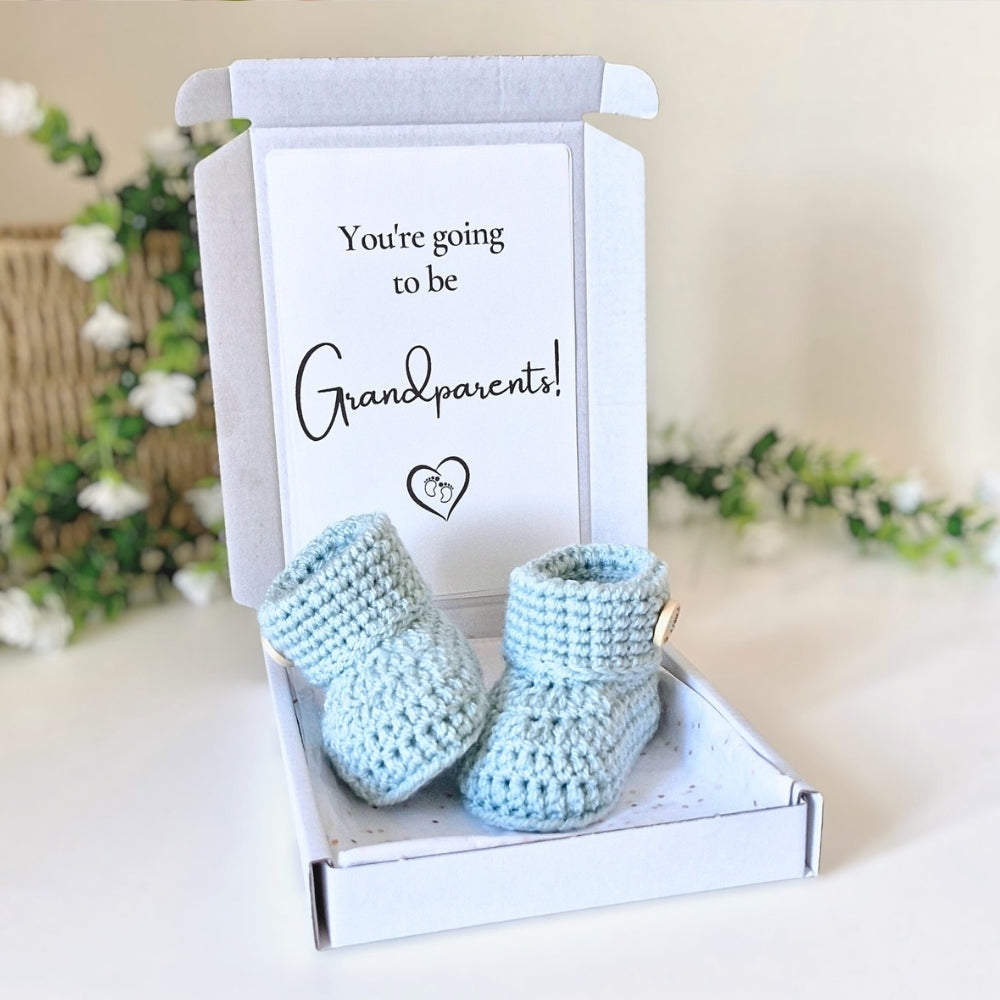 Blue baby booties in a box with a card reading 'You're going to be grandparents!' on a light background.