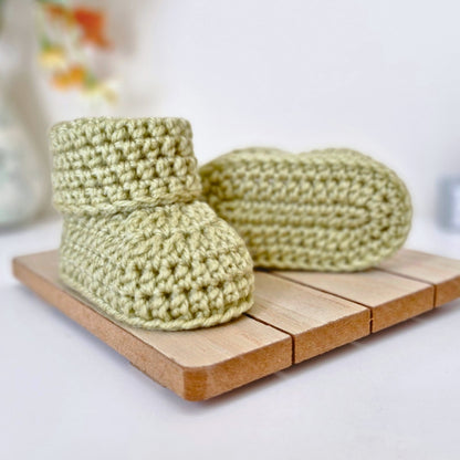 A Pair of newborn mushy peas green crochet baby booties on a wooden surface. Made with acrylic yarn