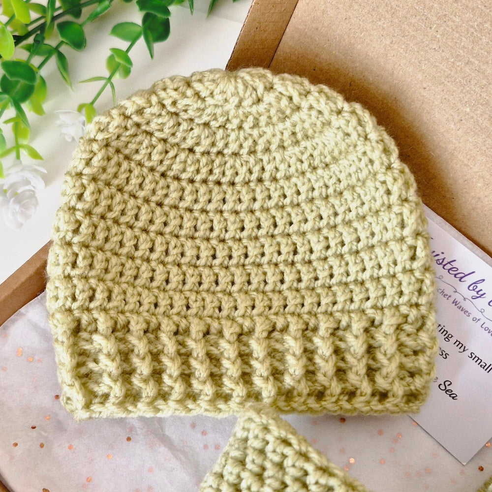 A handmade pale yellow-green crocheted baby beanie hat with a ribbed brim, displayed on a white surface with a brown cardboard box, green leaves, and a small card.