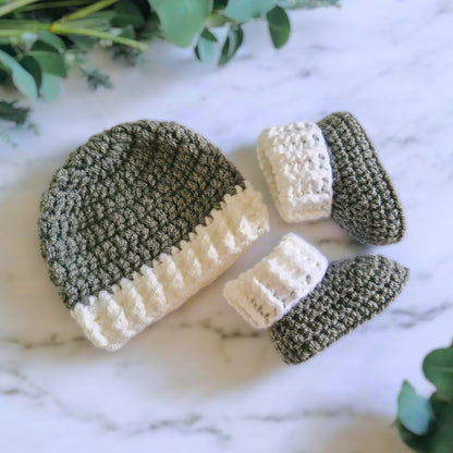 Crocheted baby hat and booties set on a marble surface with greenery.
