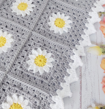 Silver crocheted blanket with white daisy flowers and yellow centers on a wooden surface.