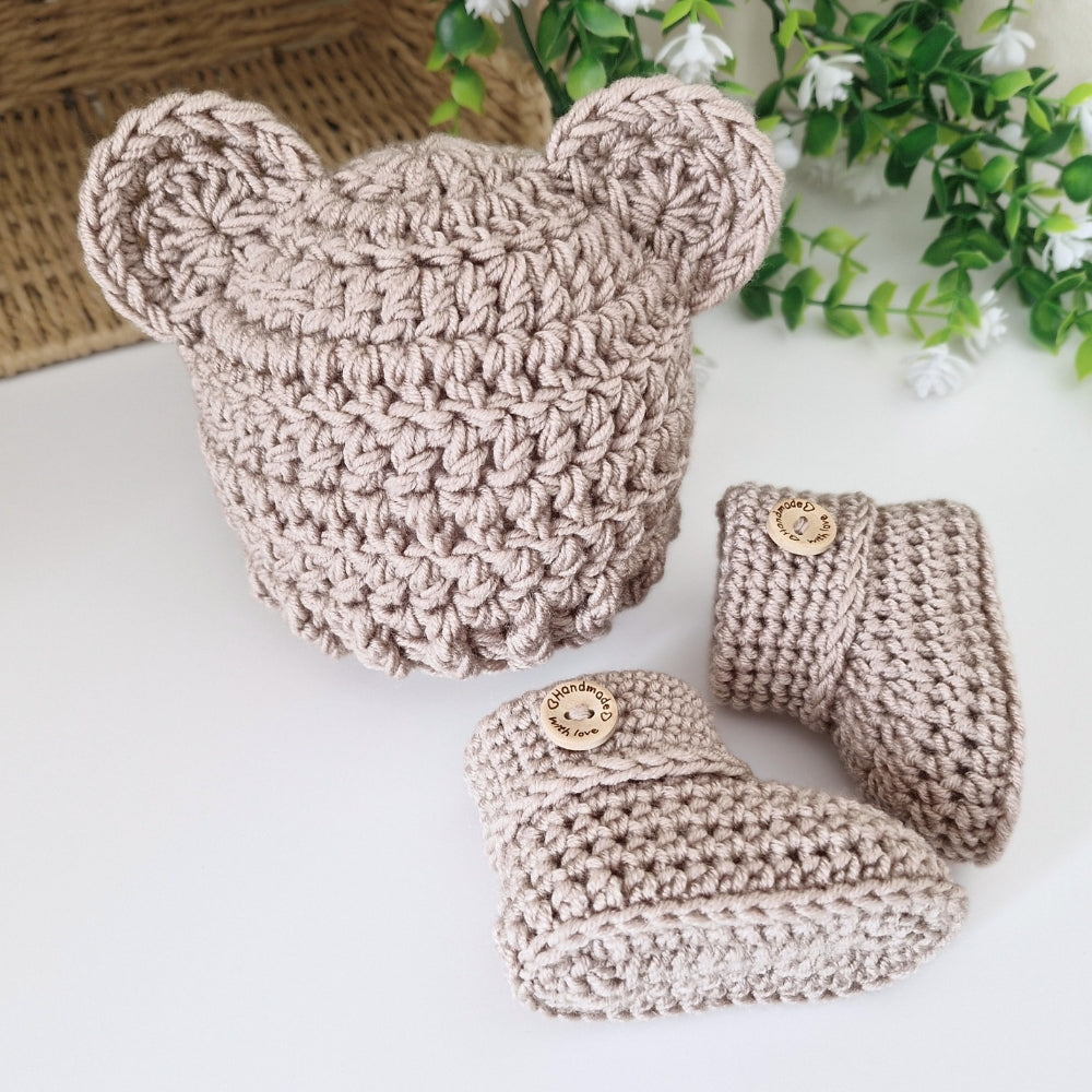 A handmade crochet baby bootie set in a warm taupe-beige color, featuring a soft crocheted hat with bear ears and matching booties with decorative wooden buttons, 