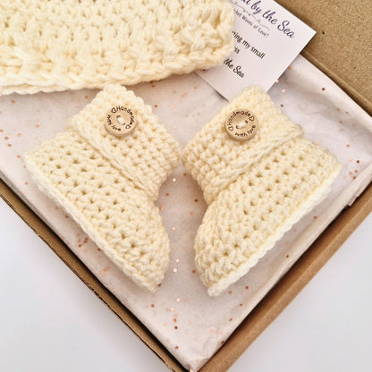 A set of cream-colored handmade crochet baby booties and a matching hat displayed in a gift box with glitter tissue paper and a brand card.