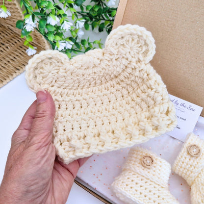 A hand holds a cream-colored crocheted baby hat with bear ears, with matching crocheted booties and a kraft gift box in the background.