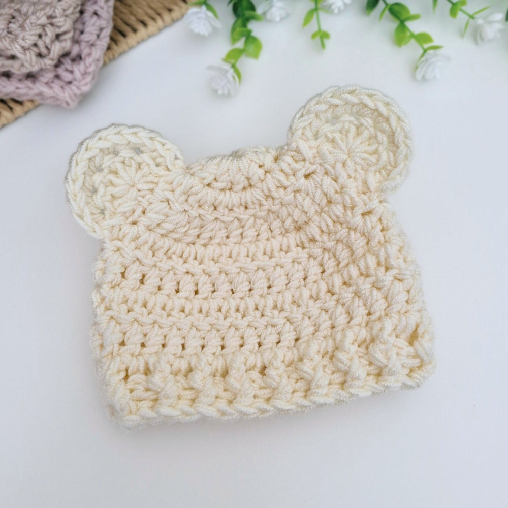 A cream-colored crocheted baby hat shaped like a bear head, displayed on a clean white surface with decorative flowers and another crocheted item in the background.